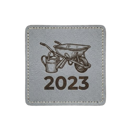 Gray square patch with stitched border. Features a detailed illustration of a wheel barrel and watering can with text "2023".